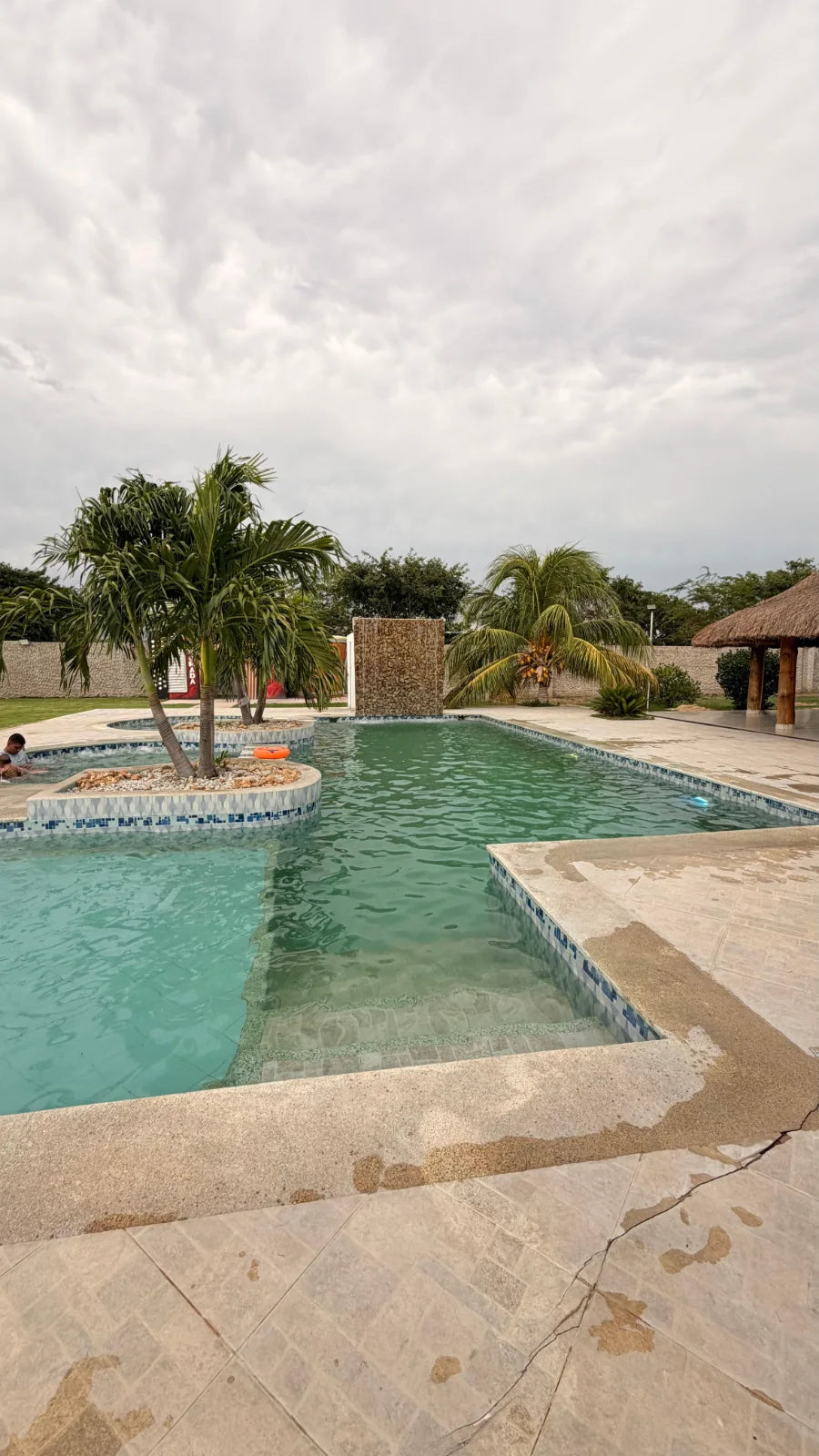 Piscina con escalones, isla de palmeras y kiosco en el lateral.