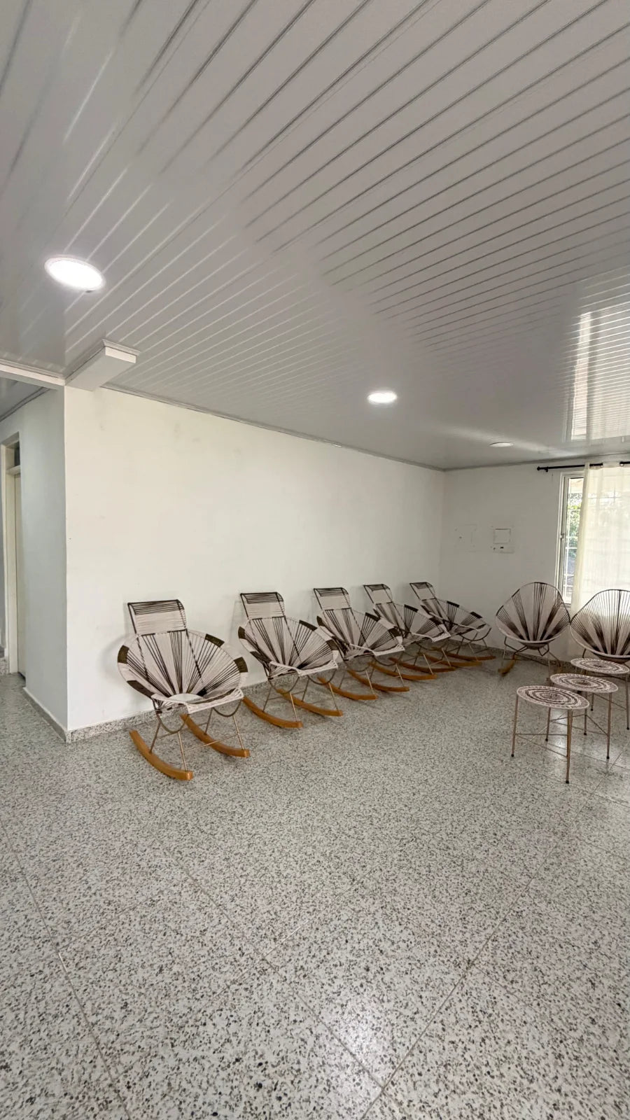 Sala interior con mecedoras, sillas tejidas y pequeñas mesas redondas.