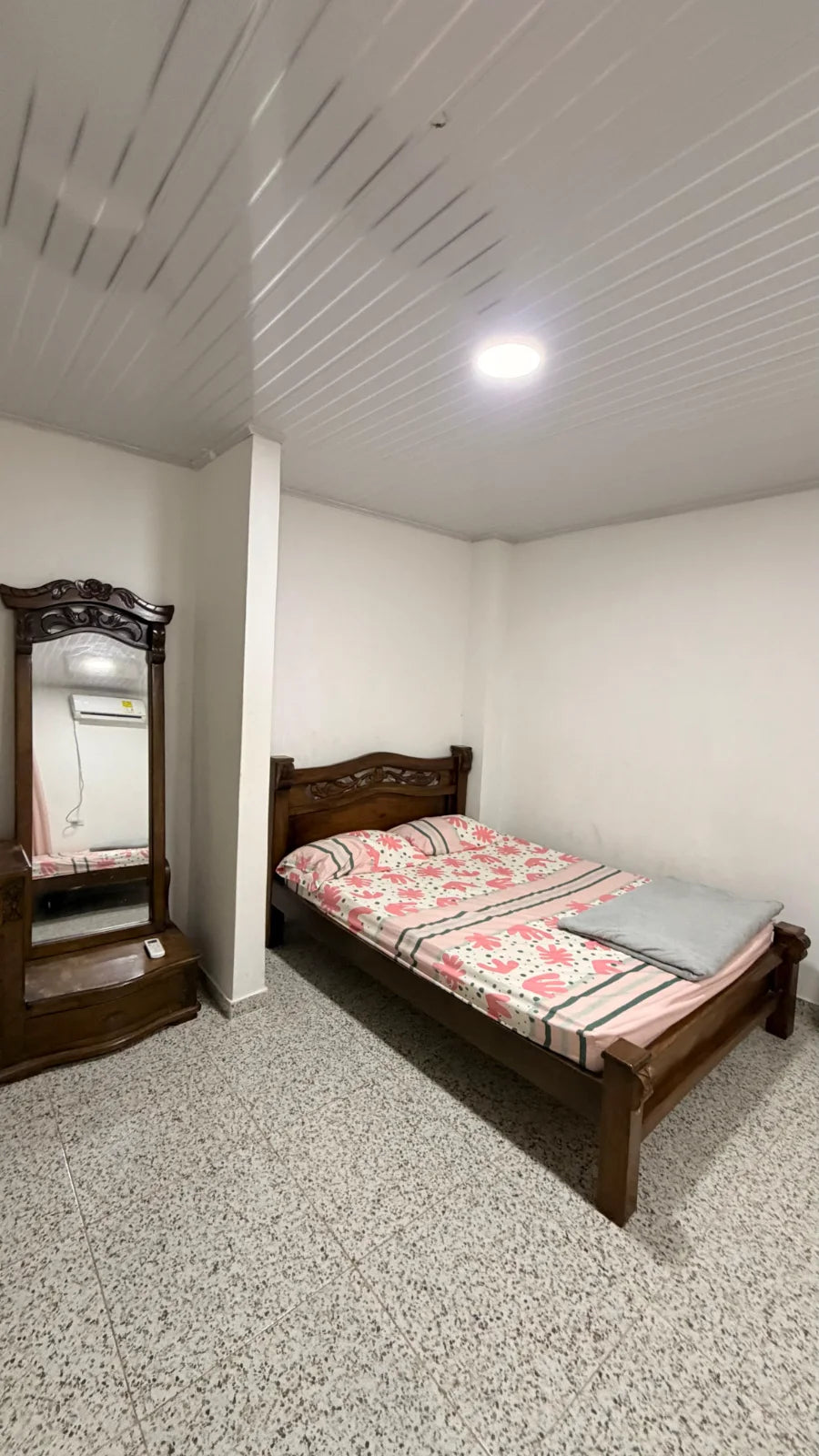 Cama de madera con ropa rosa junto a un tocador y espejo de cuerpo entero.
