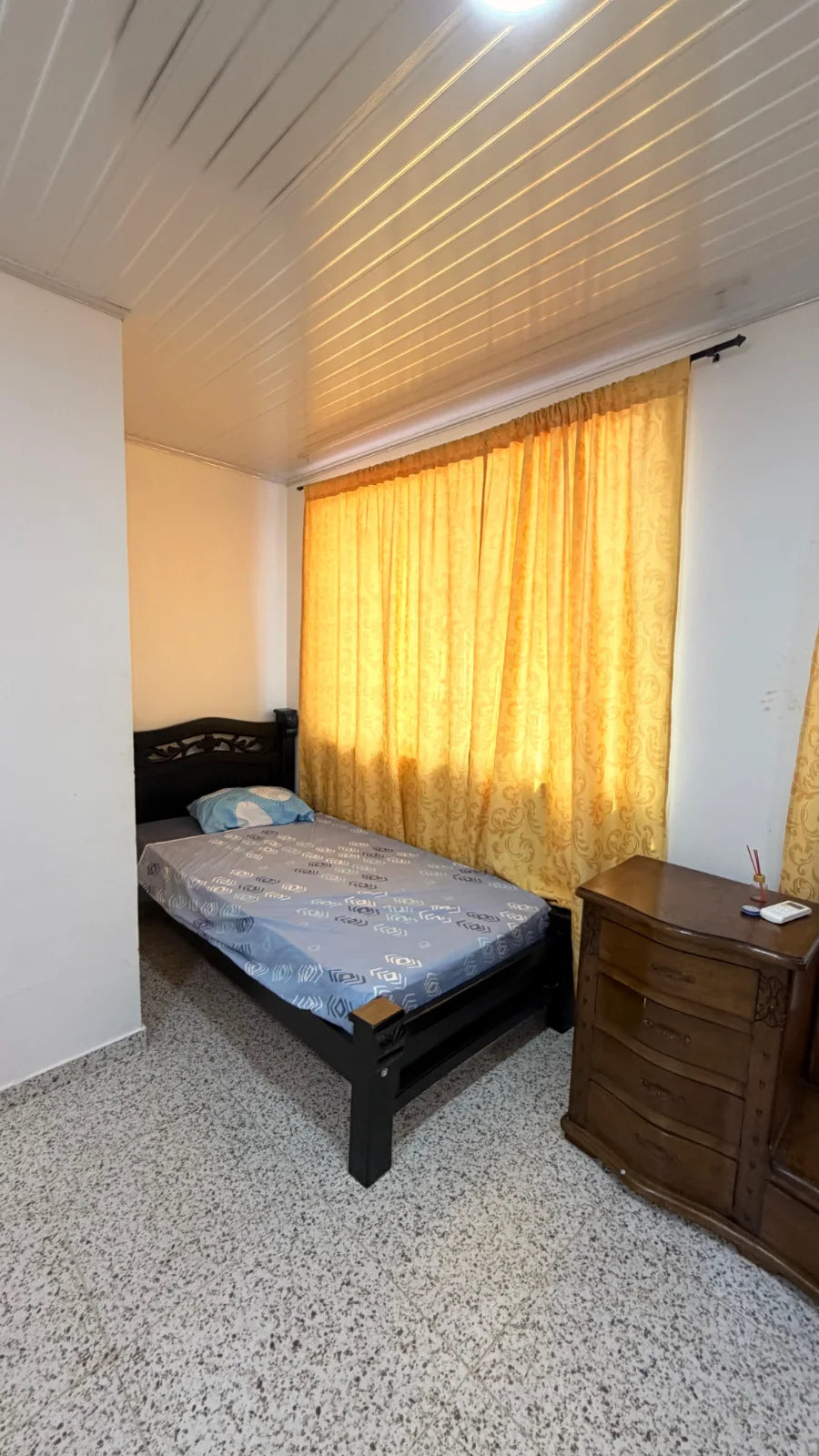 Cama de madera negra junto a una cortina amarilla y una cajonera de madera.
