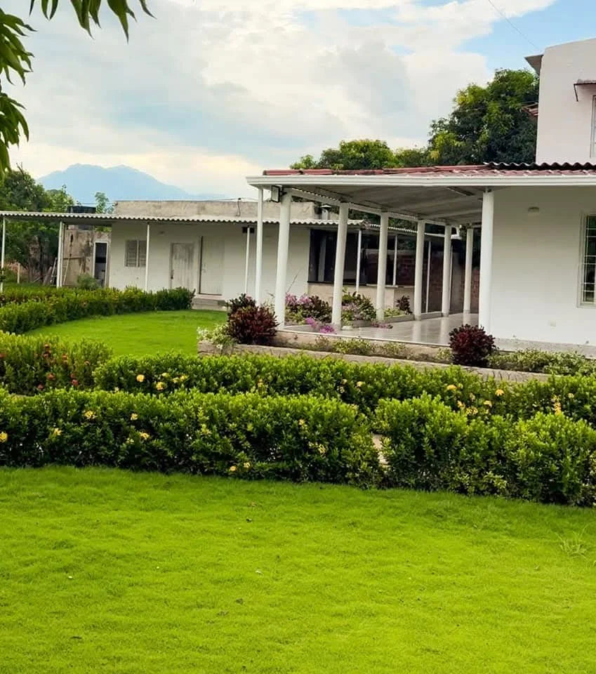Césped y setos frente al corredor blanco de la casa y un anexo bajo.